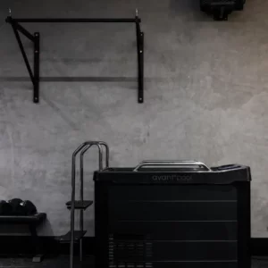 A black Avantopool Kinos Plus with metal steps is placed against a grey wall, beside a mounted pull-up bar and a bench stocked with towels.