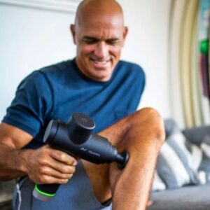 A man sits on a sofa, smiling as he uses the Hypervolt 2 massage gun on his thigh. He is wearing a dark t-shirt and shorts.