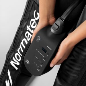 A person holds the Normatec 3, attached to leg compression sleeves, with control buttons and a digital display visible.