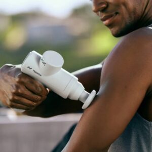 A person uses the Hypervolt GO 2 handheld massage gun on their upper arm while wearing a sleeveless top outside.