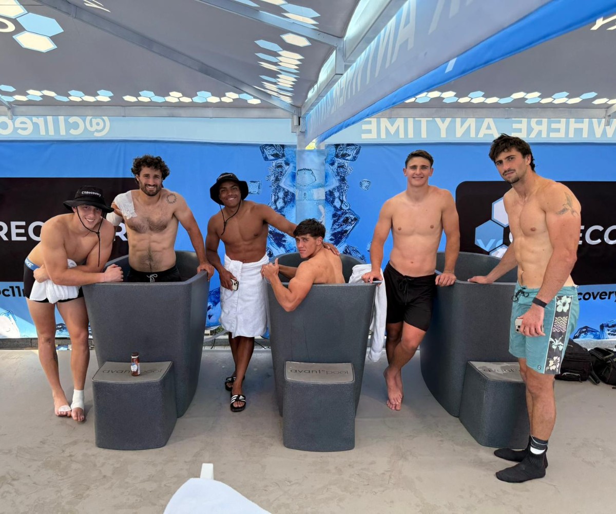 Six young men in swimwear stand under a shaded area, with three of them sitting in ice baths and three standing beside them. Fitness equipment and a drinks bottle are visible.