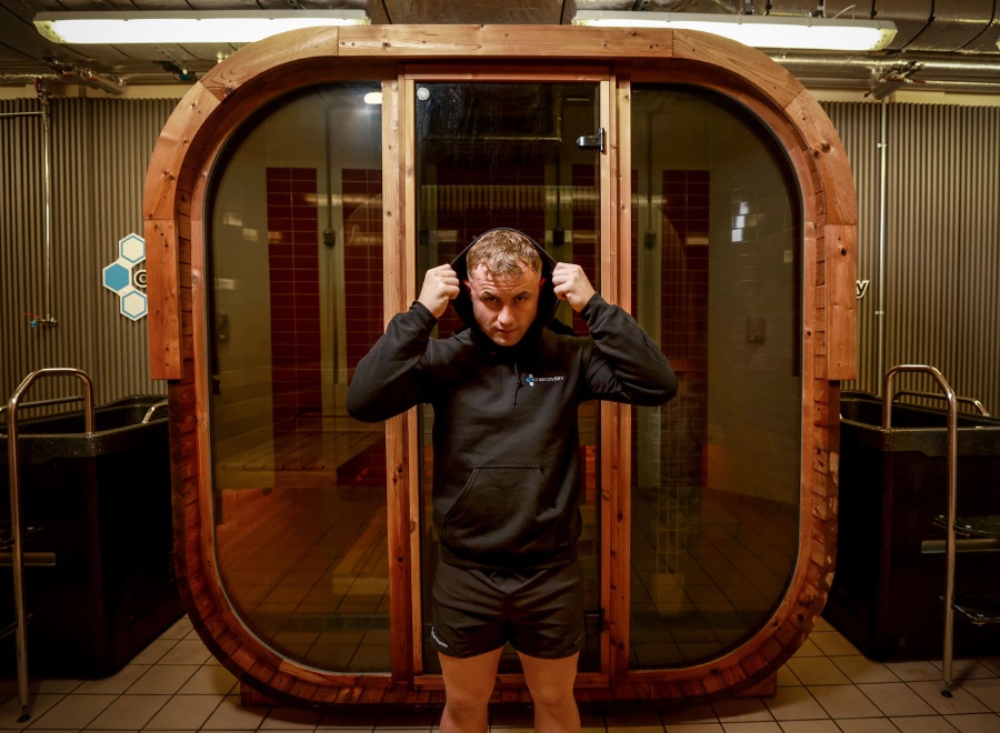 A man in a black hoodie and shorts stands indoors, adjusting his hood in front of a large, oval-shaped wooden sauna.