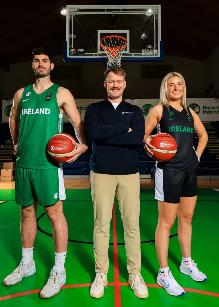 Two basketball players in Ireland uniforms stand on either side of a man in casual attire, all posing on an indoor basketball court in front of a hoop.