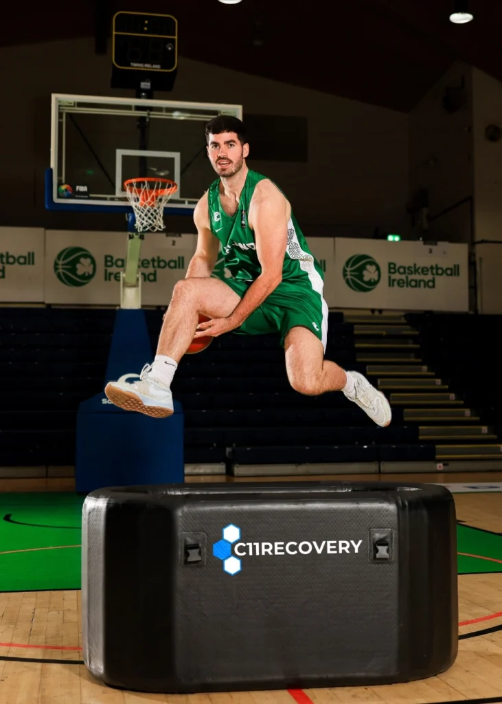A basketball player in a green kit jumps over a black C1 Recovery block on an indoor court, with a basketball hoop and Basketball Ireland signs in the background.