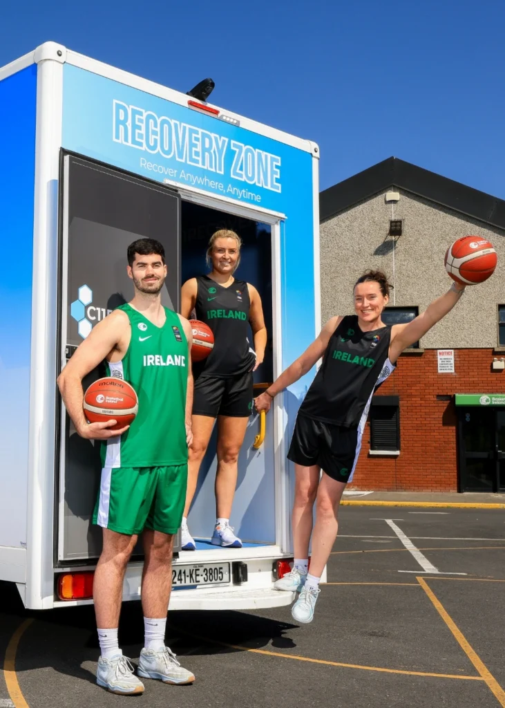 Three basketball players in Ireland kits pose with basketballs outside a mobile Recovery Zone unit in a car park on a sunny day.