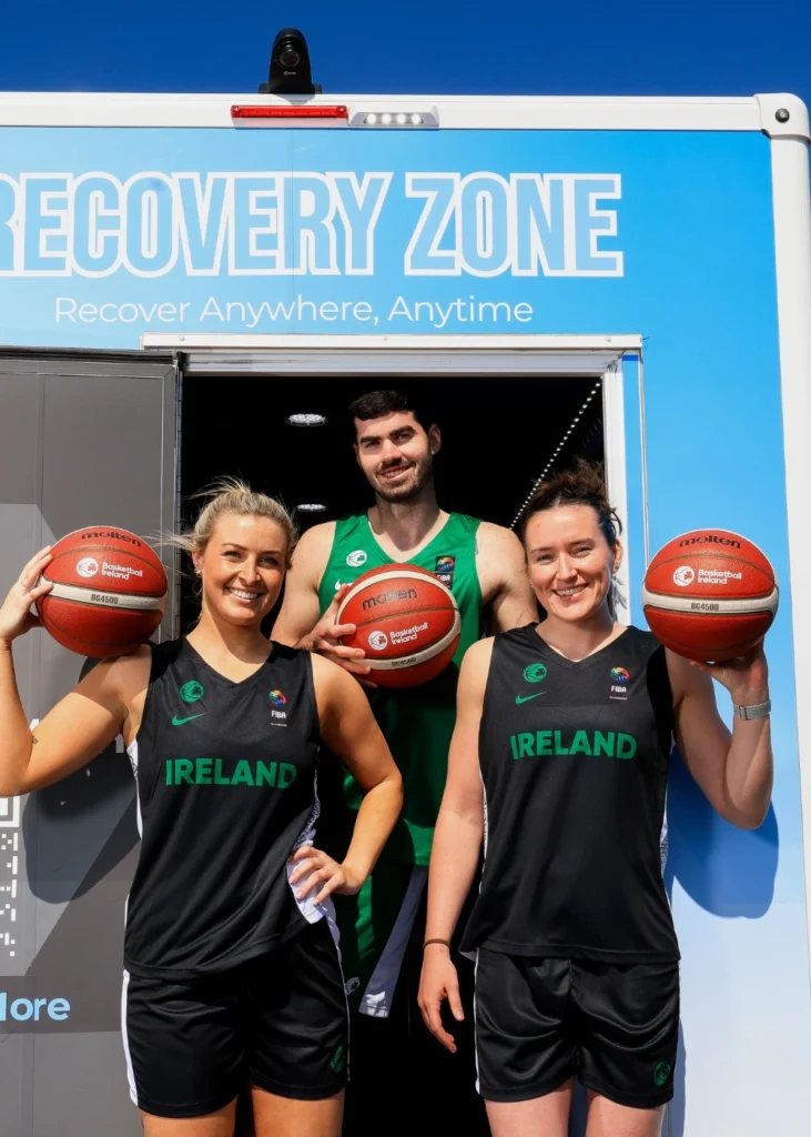 Three basketball players in Ireland jerseys stand in front of a Recovery Zone trailer, each holding a basketball and smiling at the camera.