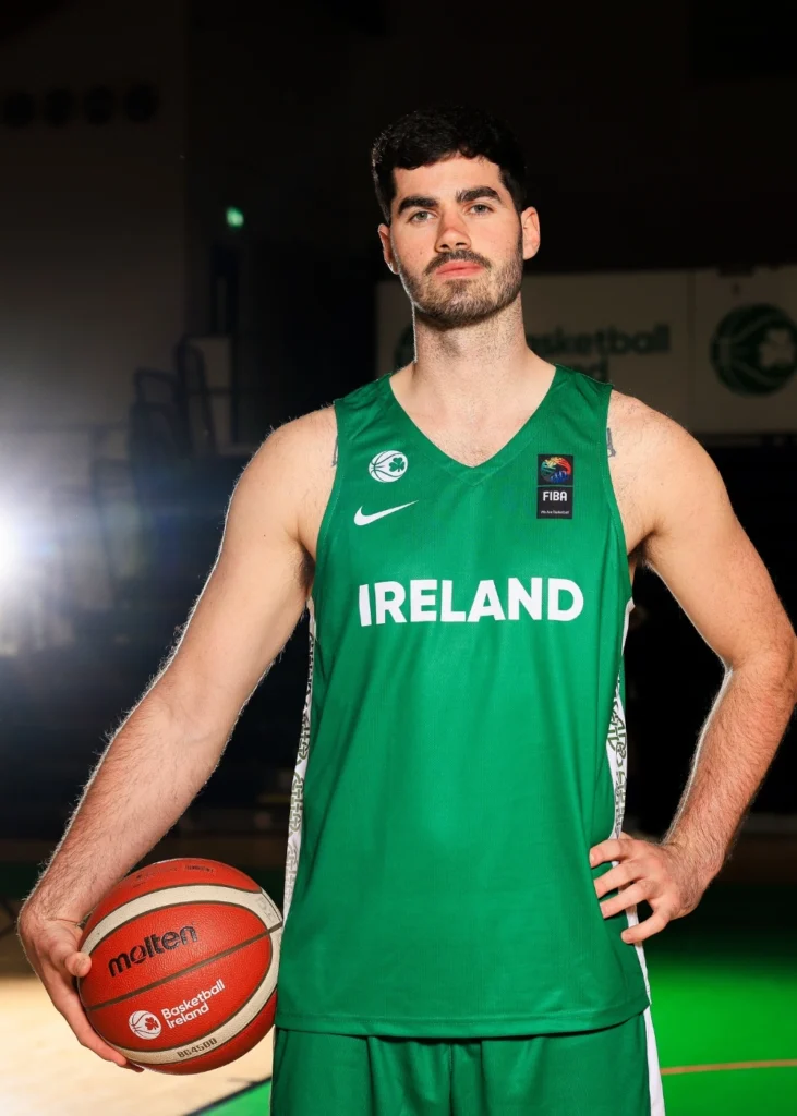A basketball player in a green Ireland jersey stands holding a basketball in his right hand, posing on an indoor court with lights in the background.