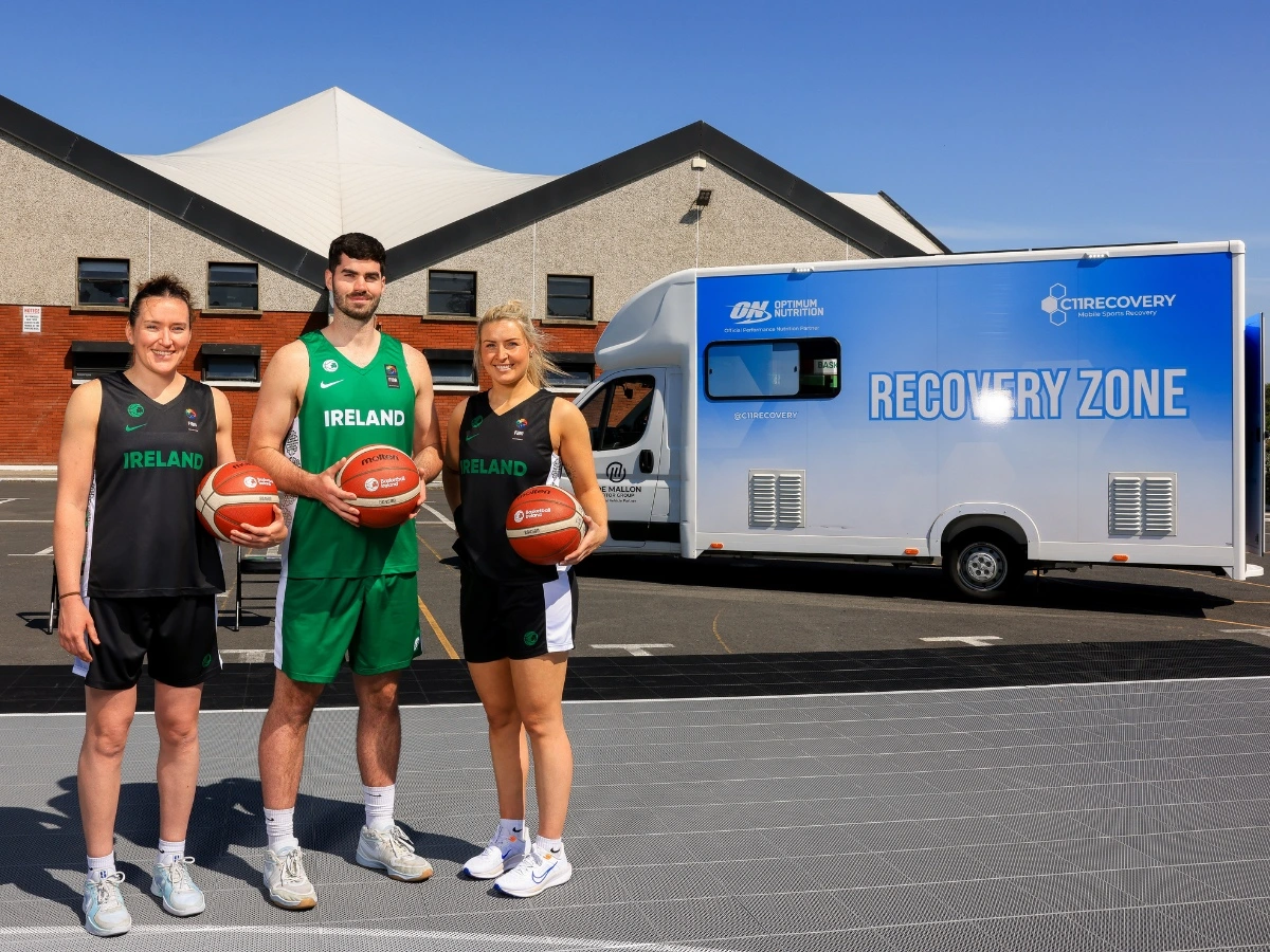 Three basketball players in Ireland kit pose with basketballs on an outdoor court, standing in front of a blue Recovery Zone van under a clear sky—showcasing how the National Team Recovery helps them recover like the pros.