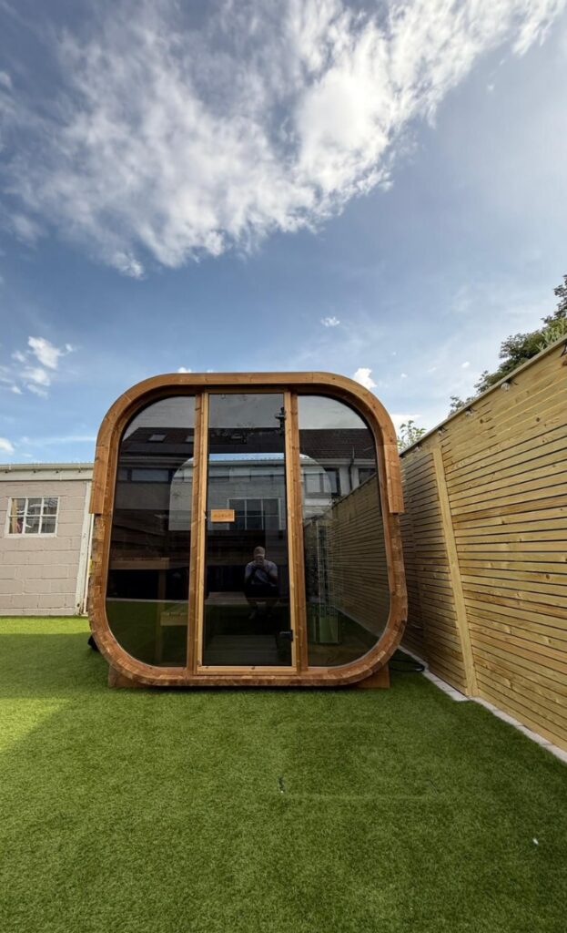 A small, modern wooden garden pod with large glass doors, perfect as a cosy sauna, sits on artificial grass, surrounded by a wooden fence and buildings under a partly cloudy sky.