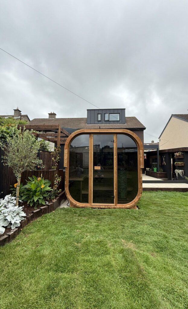 A modern, square-shaped wooden garden pod with large glass doors, perfect for use as a sauna, is situated on a green lawn with houses and an overcast sky in the background.