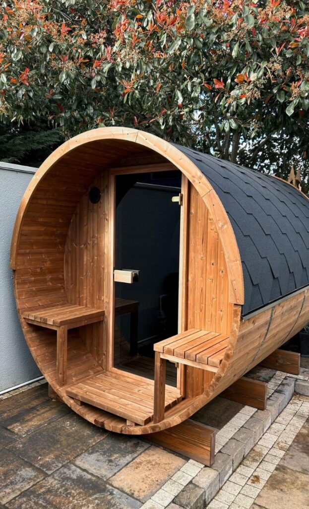 A barrel-shaped wooden outdoor sauna with a tiled roof, benches at the entrance, and stone paving in a garden setting.