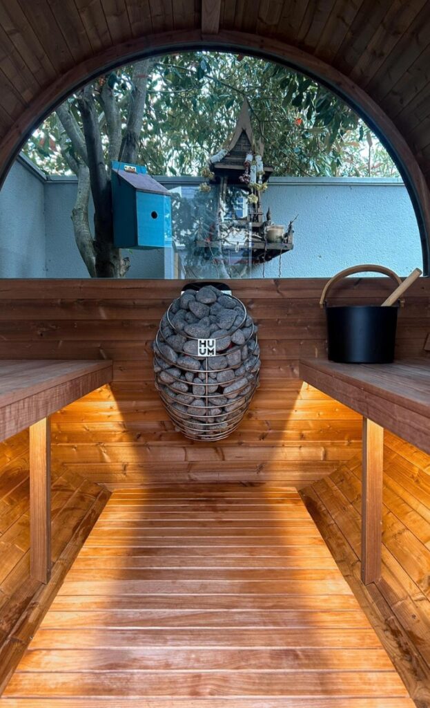 Wooden sauna interior with a metal stove filled with stones at the centre, benches on both sides, and a large window showing a bird box and tree outside.