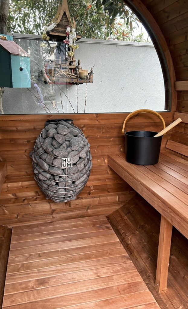 Interior of a wooden sauna with a metal basket of stones attached to the wall and a black bucket with a wooden ladle on the bench.