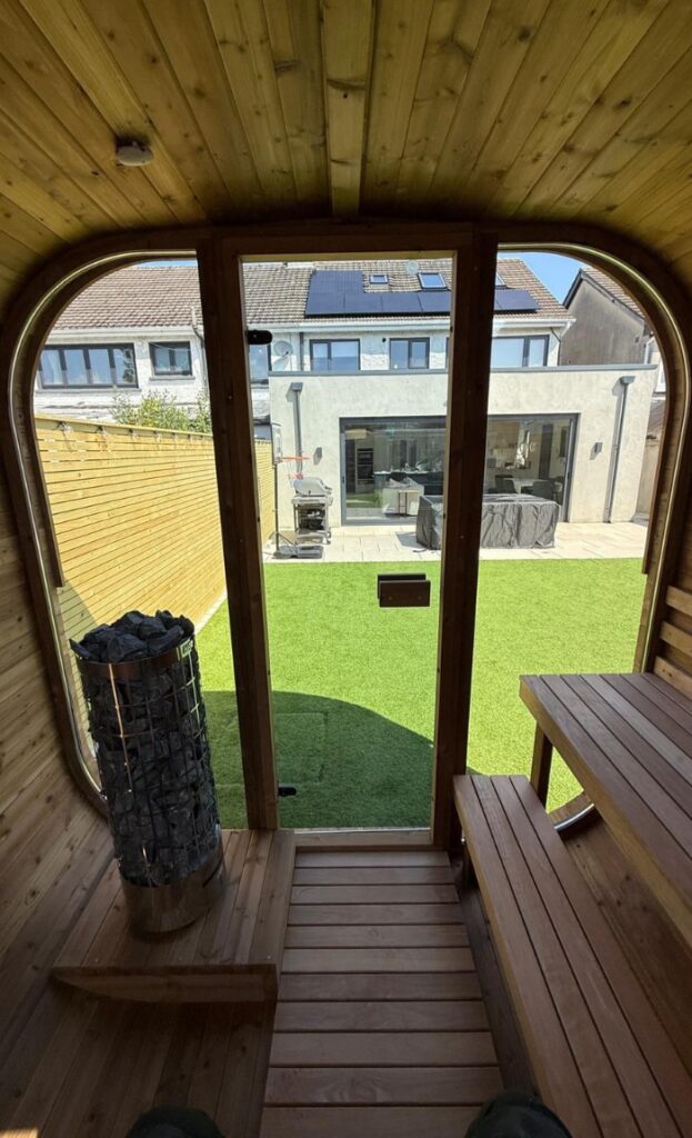 View from inside a cosy wooden sauna looking out through a glass door at a back garden featuring artificial grass, a barbecue, and a modern house.
