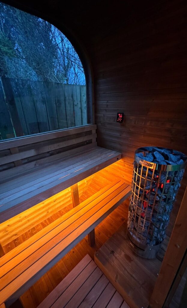 Interior of a wooden sauna with lit benches and a metal sauna heater filled with stones, next to a window showing an outdoor fence and trees at twilight.