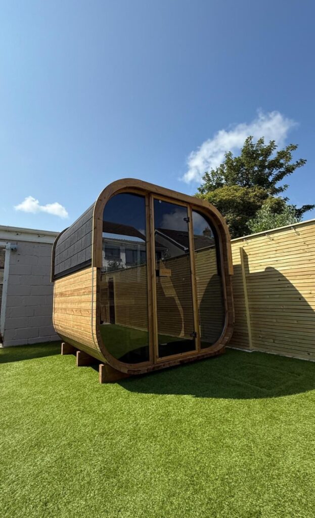 A modern wooden garden pod with large glass doors, perfect for use as a sauna, sits on artificial grass, enclosed by a wooden fence under a clear blue sky.