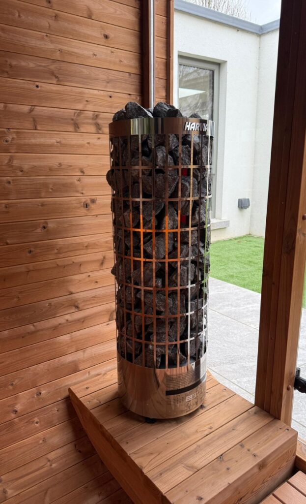 A modern electric sauna heater filled with dark stones is positioned on a wooden platform inside a wooden-panelled sauna room with a view of an outdoor area.