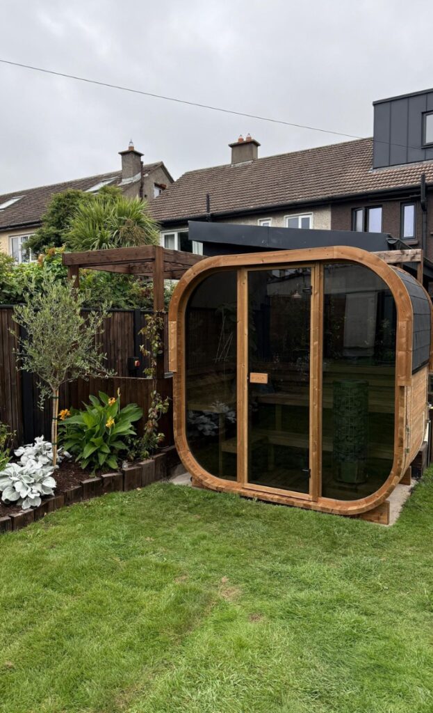 A wooden barrel-shaped sauna with large glass doors sits on a lawn, surrounded by plants and a wooden fence, with houses in the background under a cloudy sky.