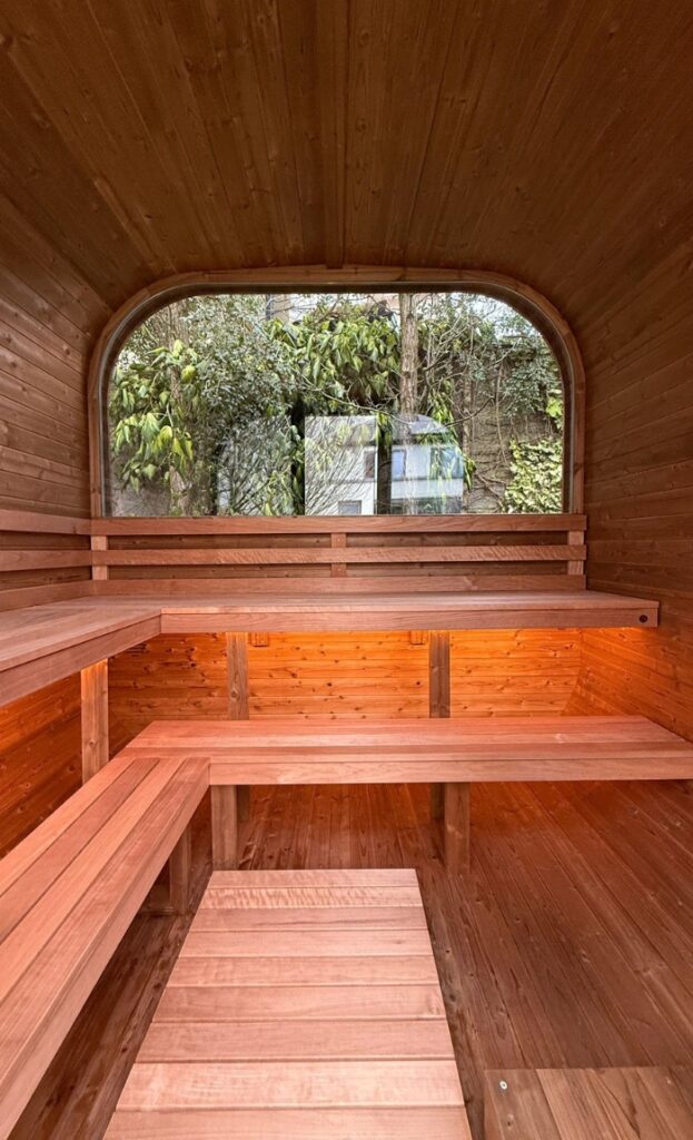 Interior of a wooden sauna with tiered seating and a large window offering views of lush greenery outside, creating a relaxing sauna haven.