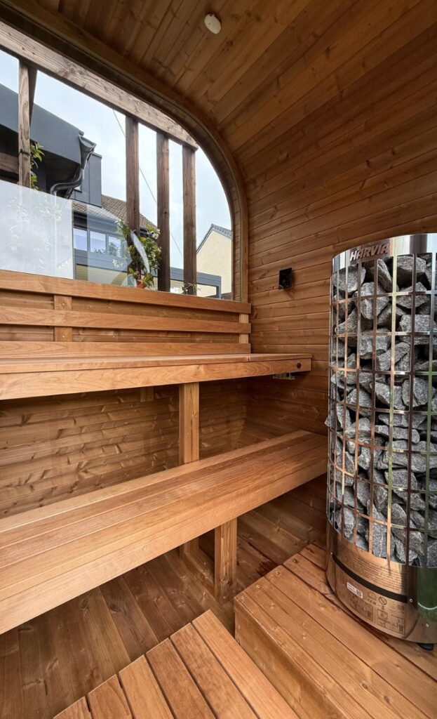 Interior of a wooden sauna featuring tiered benches and a metal electric sauna heater filled with stones; window reveals an inviting outdoor view.