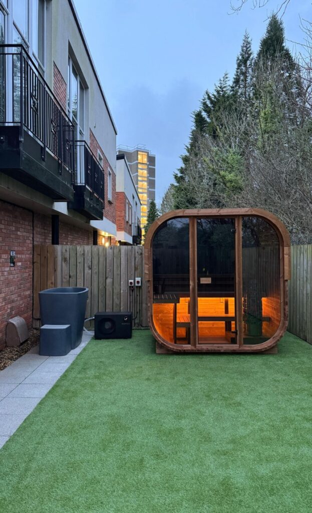 A modern garden featuring artificial grass, patio seating, a cosy wooden sauna with interior lights, a speaker, and a wooden fence, all nestled between blocks of flats and lush trees.