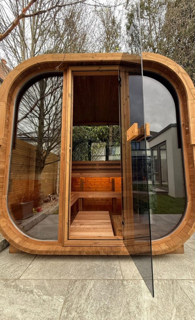 A modern wooden outdoor sauna with a large glass door, interior benches, and ambient lighting sits on a tiled patio surrounded by trees and a house, offering the perfect place to unwind in nature.