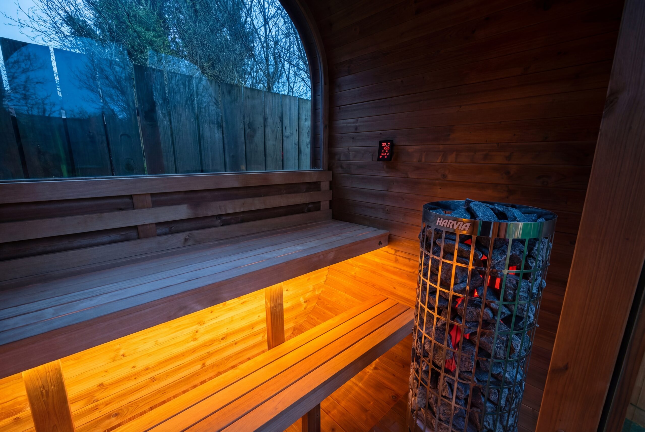 A wooden sauna interior with benches, a basket of sauna stones heater, and warm yellow lighting; a window shows an outdoor view.