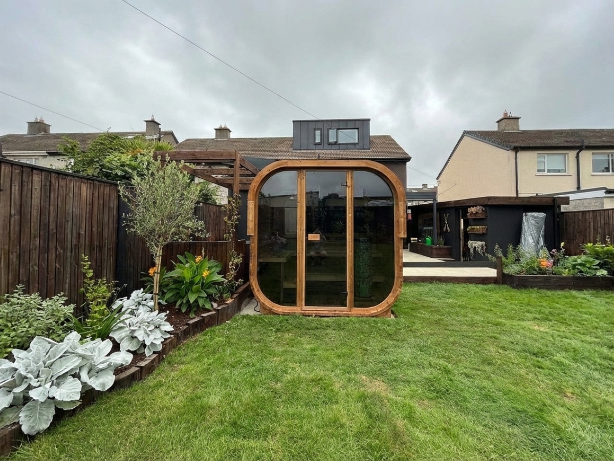 A modern, timber-framed garden room with large glass doors sits on a lawn in a fenced back garden, surrounded by various plants and shrubs.