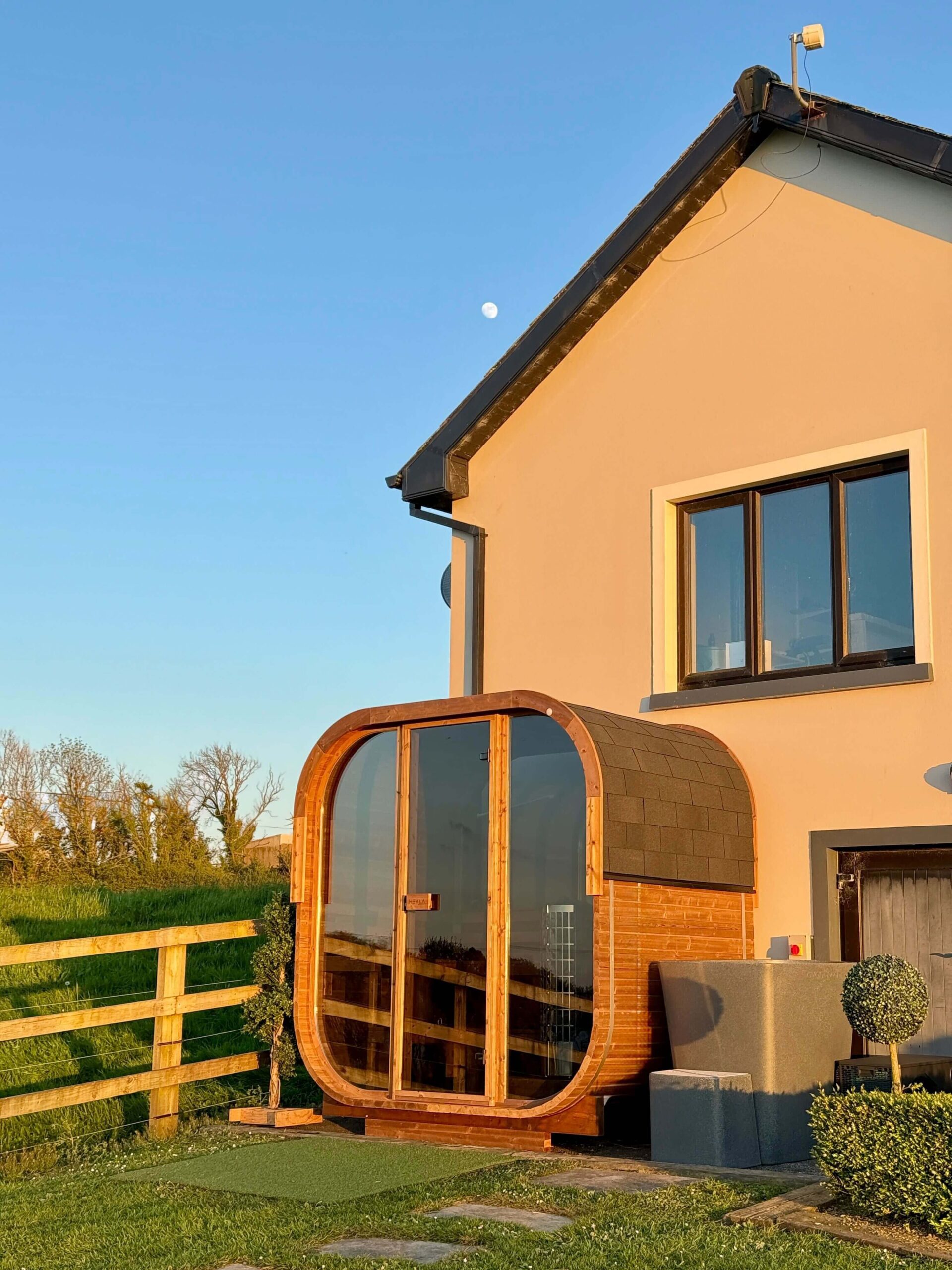 A small wooden sauna, perfect for relaxing between ice baths, sits beside a cream-coloured house, with a fence and grassy garden in the background under a clear evening sky.