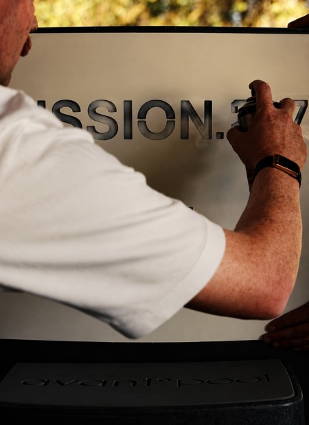 Person in a white shirt spray paints stencilled letters onto a surface, with part of the text MISSION.37 visible.