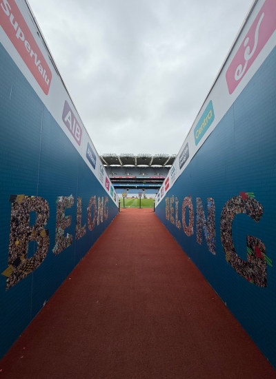 View from a stadium tunnel with BELONG spelt out on the walls, leading to a pitch under a cloudy sky—capturing the passion and excitement of Irish sport during All-Ireland Final Weekend.