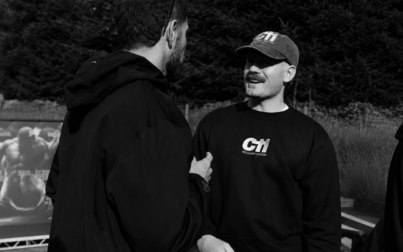 Two men in casual clothing talk outdoors. One wears a cap and sweatshirt with CH1 branding. Trees and a blurred billboard are visible in the background.