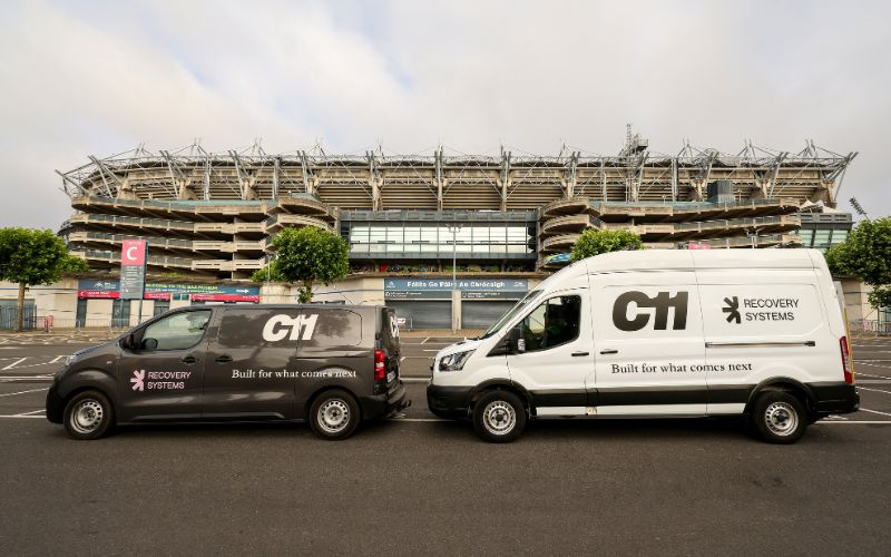 A dark grey van and a white van, both branded with C11 Recovery Systems, are parked side-by-side in a large car park in front of Croke Park stadium during All-Ireland Final Weekend, when the excitement of Gaelic football fills the air.
