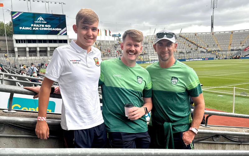 Three men standing and smiling in a sports stadium during the All-Ireland Final Weekend, with empty seats and a scoreboard visible in the background.