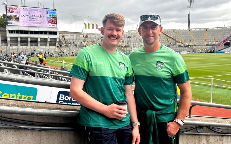 Two men in matching green sports shirts stand together in a stadium near the pitch, with empty seats and a scoreboard visible in the background, capturing the anticipation of All-Ireland Final Weekend.