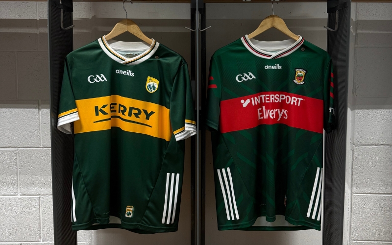 Two green GAA jerseys hang on wooden hangers: a Kerry jersey with a yellow stripe and a Mayo jersey with a red stripe, displayed against a white brick wall—perfect for showing your county pride ahead of All-Ireland Final Weekend.