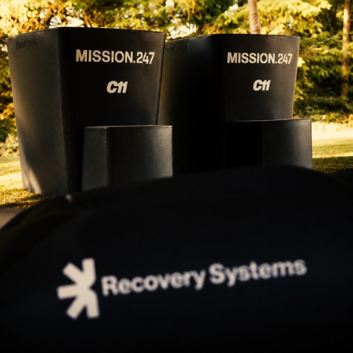 Two black containers labelled MISSION.24/7 C11 outdoors, with a blurred Recovery Systems logo on a foreground object. Trees in the background.