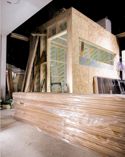 A partially built plywood room with a large stack of timber planks wrapped in plastic in the foreground and construction materials around it.