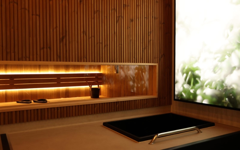 A cold plunge set into a beige tiled surround with a stainless handrail, in front of a wooden slat wall with a backlit shelf and a bright window.