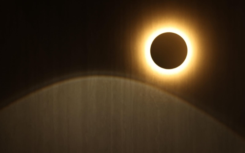 A round backlit wall light glowing like an eclipse above a curved timber panel in a dark room at New Era.