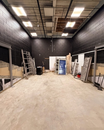 A large empty room with black walls and an exposed ceiling, with ladders, planks, a door and construction materials lined along the walls.