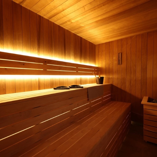 A wooden sauna room with built-in bench seating, indirect lighting, a bucket with a ladle and towels placed on the bench.