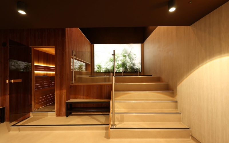 Beige tiered steps with a glass balustrade, a wood-panelled sauna on the left and a large window with greenery outside, lit with warm ambient lighting.