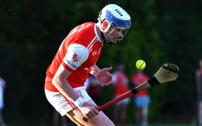 A hurling player in a red and white kit prepares to strike a sliotar with a hurley during a match, with blurred players in the background.