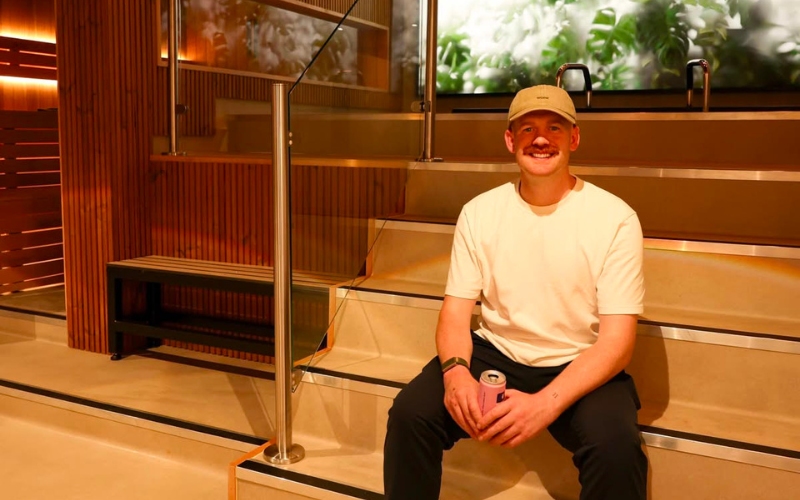 Ryan Casey in a white t-shirt and beige cap sitting smiling on the tiered steps of the finished New Era recovery room, with timber slat walls and greenery behind him.
